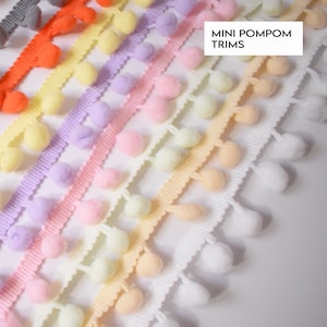 May include: A selection of mini pompom trims in a range of pastel shades: grey, orange, yellow, lilac, pink, and white. Each trim features a scalloped edge. These trims are suitable for craft and sewing projects. The text "MINI POMPOM TRIMS" is visible.