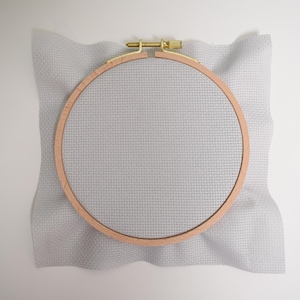 May include: A white embroidery hoop with a wooden frame and a light gray Aida cloth.