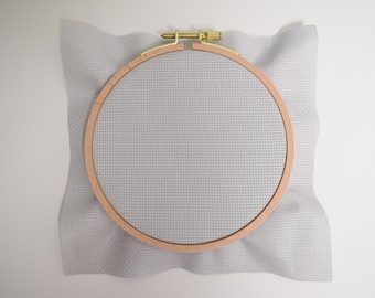 May include: A white embroidery hoop with a wooden frame and a light grey Aida cloth.