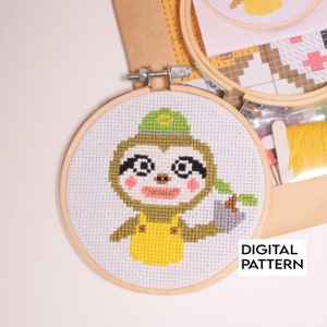 May include: Cross stitch pattern of a sloth wearing a green hat and yellow overalls, holding a bucket. The pattern is in a wooden embroidery hoop. The text "DIGITAL PATTERN" is visible below the hoop.