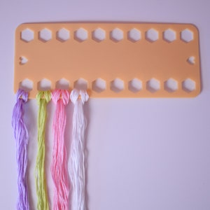 K&ouml;nnte beinhalten: Ein pfirsichfarbener Fadenorganizer mit mehreren sechseckigen L&ouml;chern und herzf&ouml;rmigen Aussparungen. Str&auml;nge farbigen Stickgarns in Lila, Gr&uuml;n, Rosa und Wei&szlig; sind befestigt, bereit f&uuml;r Bastelprojekte.