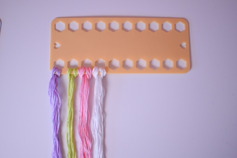 K&ouml;nnte beinhalten: Ein pfirsichfarbener Fadenorganizer mit mehreren sechseckigen L&ouml;chern und herzf&ouml;rmigen Aussparungen. Str&auml;nge farbigen Stickgarns in Lila, Gr&uuml;n, Rosa und Wei&szlig; sind befestigt, bereit f&uuml;r Bastelprojekte.