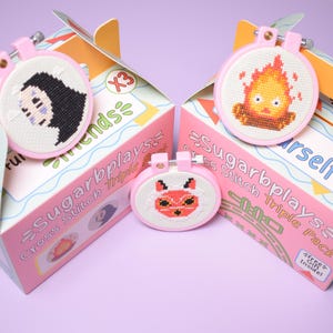 May include: Three cross-stitch kits with pink hoops and boxes. The kits feature cartoon character designs, including a person with black hair, a fiery creature, and a red fox. The boxes are pink and white with the text "Sugarbplays Cross Stitch Triple Pack".