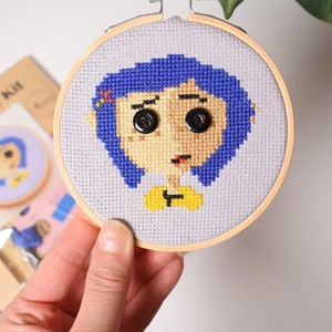 May include: A cross-stitch embroidery hoop with a colorful image of a girl with blue hair and a yellow shirt. The girl's eyes are black buttons.