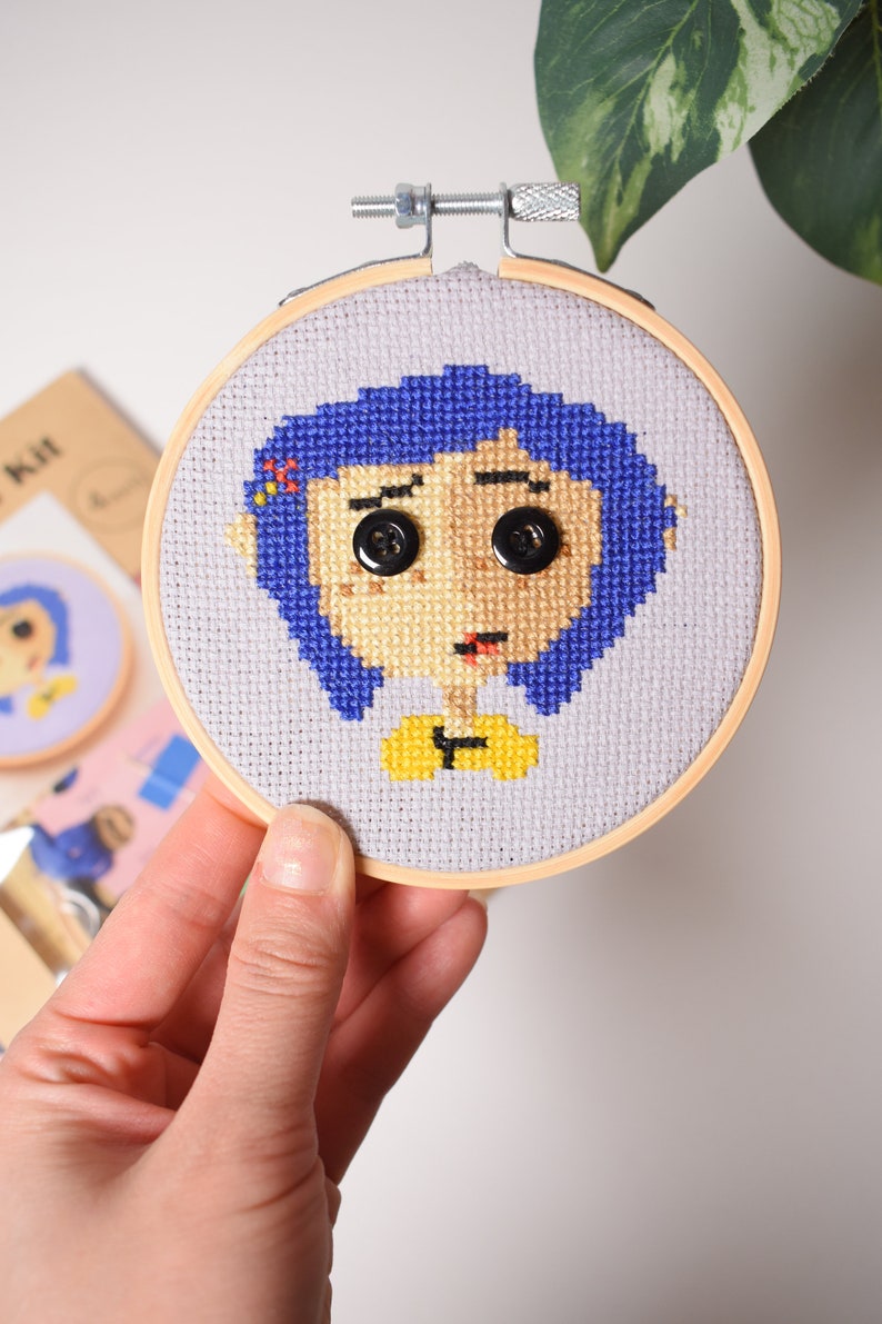 May include: A cross-stitch embroidery hoop with a colorful image of a girl with blue hair and a yellow shirt. The girl's eyes are black buttons.