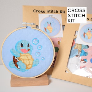 May include: Cross stitch kit featuring Squirtle from Pokemon. The kit includes a blue and yellow Squirtle design, a pre-printed Aida cloth, embroidery floss, and a wooden embroidery hoop.