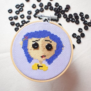 May include: A purple and blue cross-stitch embroidery of a girl with blue hair and yellow shirt. The embroidery is framed in a wooden hoop.