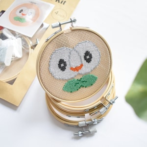 May include: A cross-stitch kit featuring a cartoon owl with green leaves. The kit includes a wooden embroidery hoop, thread, and needles. The hoop is 4 inches in diameter.