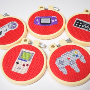 May include: Five handmade cross-stitch hoops featuring retro video game controllers and consoles. Each design is stitched on red fabric and set in a cream-colored hoop. The designs include a Game Boy, a Nintendo controller, and a Nintendo 64 controller.