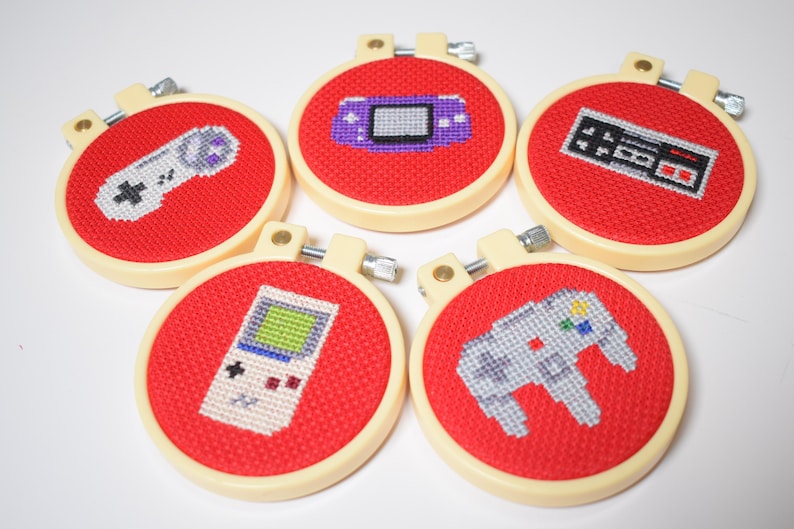 May include: Five handmade cross-stitch hoops featuring retro video game controllers and consoles. Each design is stitched on red fabric and set in a cream-colored hoop. The designs include a Game Boy, a Nintendo controller, and a Nintendo 64 controller.