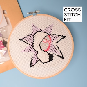 May include: A cross stitch kit featuring a pink and black design of a stylized face with a starburst behind it. The kit includes a hoop, fabric, and floss.