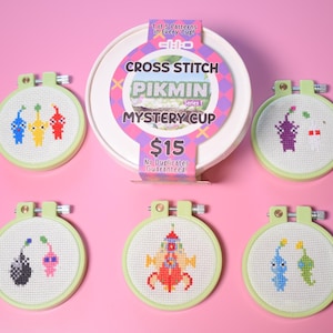 Puede incluir: Una colección de seis diseños de Pikmin en punto de cruz en aros de bordado, con un recipiente blanco en el centro. Los aros son de color verde claro y los diseños de punto de cruz presentan varios personajes Pikmin de colores. El recipiente tiene el texto "CROSS STITCH PIKMIN MYSTERY CUP".