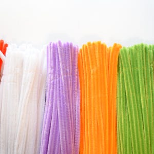 Puede incluir: Una colección de limpiapipas difusos dispuestos en haces verticales, con un espectro vibrante de colores que incluye rojo, blanco, lavanda, naranja, verde lima y azul. Cada tallo individual tiene una superficie suave, texturizada y flexible.