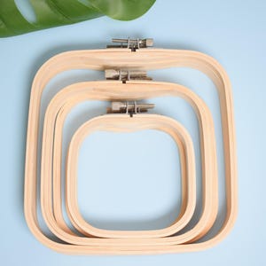 Op de afbeelding: Drie houten borduurringen van verschillende maten, in elkaar genest. De ringen zijn vierkant met afgeronde hoeken en hebben een natuurlijke houten afwerking. Metalen hardware zet de stof vast. De achtergrond is lichtblauw met groene bladeren.