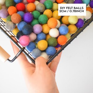 May include: A black wire basket filled with colorful felt balls, each measuring 2 cm or 0.78 inches in diameter. The basket is being held by a hand, and the text "DIY FELT BALLS 2CM / 0.78INCH" is visible in the upper right corner.