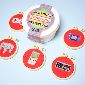 May include: A collection of red cross-stitch hoops featuring various video game controller designs. The hoops are arranged around a white container labeled "Nintendo Mystery Cup". The text "CROSS STITCH" and "Nintendo" are visible on the container.