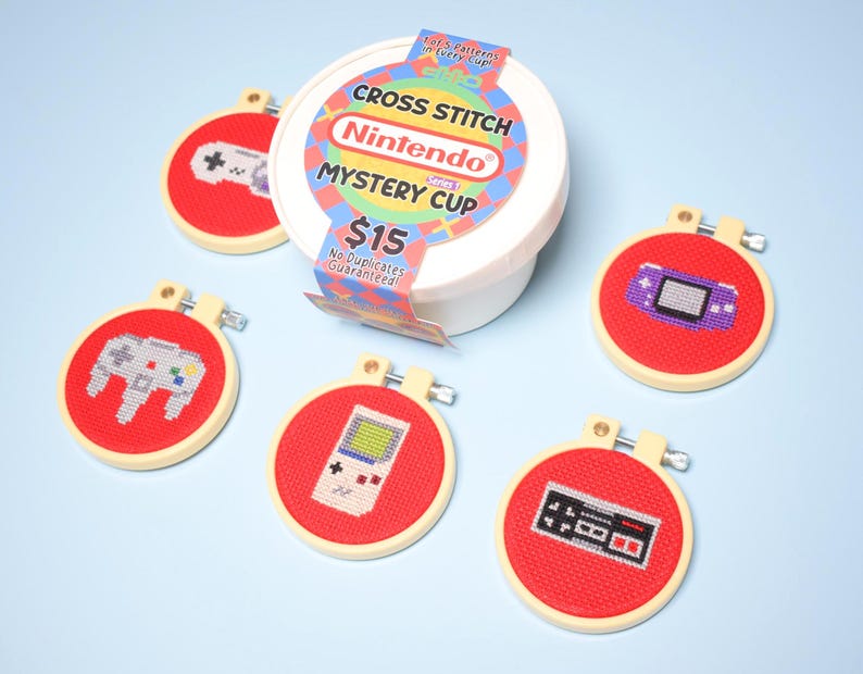 May include: A collection of red cross-stitch hoops featuring various video game controller designs. The hoops are arranged around a white container labeled "Nintendo Mystery Cup". The text "CROSS STITCH" and "Nintendo" are visible on the container.