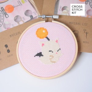 May include: A pink cross stitch kit with a white cat and a yellow orange. The kit includes a wooden hoop, thread, and a needle. The packaging says "Cross Stitch Kit" and "Material List".