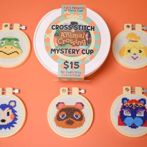 May include: A collection of six cross-stitch embroidery hoops featuring characters from Animal Crossing. The hoops are arranged around a white mystery cup with the text "Cross Stitch Animal Crossing Mystery Cup". The background is orange.