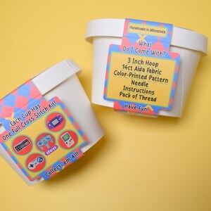 May include: Two white containers with cross-stitch kits. Each kit includes a 3-inch hoop, 14ct Aida fabric, a color-printed pattern, a needle, instructions, and a pack of thread. The kits feature retro video game graphics and the text "Collect 'em All!"