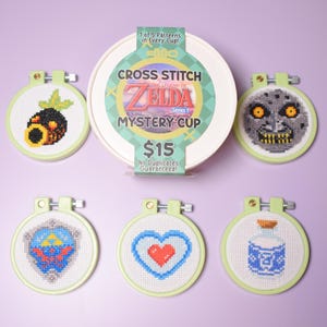 May include: A collection of cross-stitch embroidery hoops featuring designs from The Legend of Zelda series. The set includes a mystery cup with the text "CROSS STITCH THE LEGEND OF ZELDA MYSTERY CUP" and the text "1 of 5 Patterns in Every Cup!"