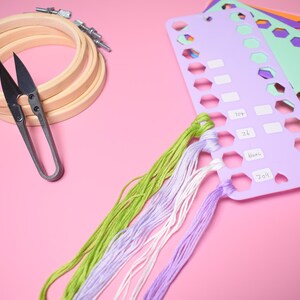 K&ouml;nnte beinhalten: Stickzubeh&ouml;r auf rosa Oberfl&auml;che. Enth&auml;lt h&ouml;lzerne Stickrahmen, eine Schere, farbenfrohes Stickgarn und Garnorganisatoren. Die Garnfarben sind Gr&uuml;n, Lila und Wei&szlig;.