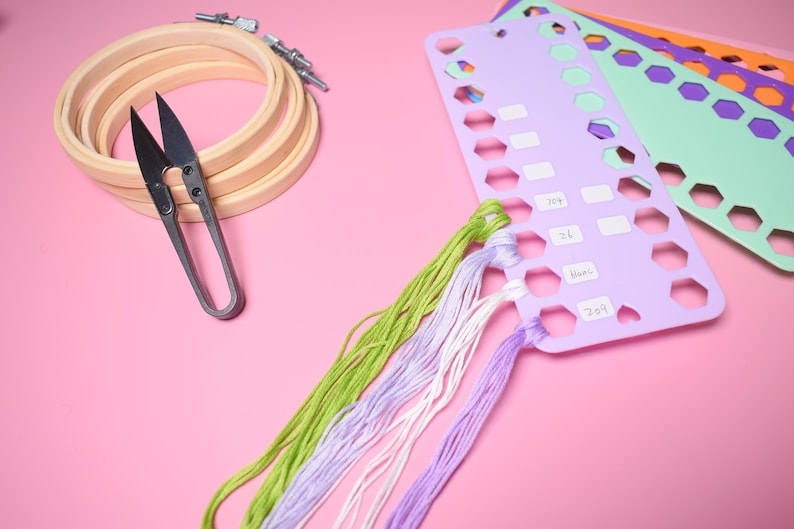 K&ouml;nnte beinhalten: Stickzubeh&ouml;r auf rosa Oberfl&auml;che. Enth&auml;lt h&ouml;lzerne Stickrahmen, eine Schere, farbenfrohes Stickgarn und Garnorganisatoren. Die Garnfarben sind Gr&uuml;n, Lila und Wei&szlig;.