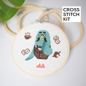 May include: A cross stitch kit featuring a teal bird with a brown satchel, surrounded by coffee-themed icons. The kit includes a wooden embroidery hoop and the text "CROSS STITCH KIT" in a white box.