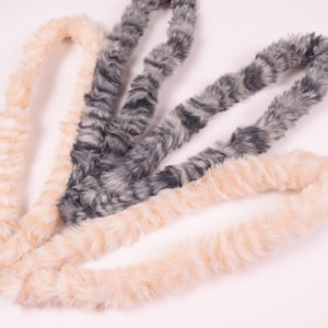 Könnte beinhalten: Nahaufnahme von drei Kunstfell-Boas in neutralen Farben. Zwei sind hellbeige und eine ist eine Mischung aus Grau und Schwarz. Die Boas sind in einem Schleifenmuster vor einem weißen Hintergrund angeordnet. Diese Accessoires eignen sich für Kostüme oder Mode.