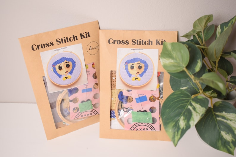 May include: Two brown paper cross stitch kits with a 4 inch hoop featuring a cartoon character with blue hair and a yellow shirt. The kits include embroidery floss, needles, and aida cloth.