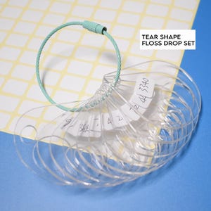 May include: A clear, tear-shaped floss drop set with a mint green metal ring. The set includes multiple clear plastic tags with handwritten numbers and text. The text "TEAR SHAPE FLOSS DROP SET" is in a white rectangle.