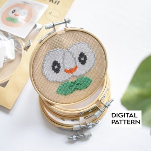 May include: A close-up of a cross-stitch pattern of a Pokemon character with large eyes and a green leaf. The pattern is in a wooden embroidery hoop. The text "DIGITAL PATTERN" is visible in the lower right corner of the image.