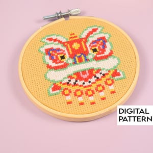 May include: A cross-stitch embroidery of a colorful lion dance head in a wooden hoop. The design features red, white, yellow, and green details on a tan background. The text "DIGITAL PATTERN" is in a white rectangle.