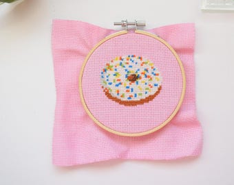 May include: A cross-stitch embroidery of a donut with white frosting and colorful sprinkles, set in a wooden hoop. The hoop is on a pink fabric background, creating a craft-themed image.