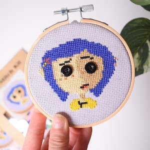 May include: A cross-stitch embroidery hoop with a purple and blue character with a yellow shirt and black button eyes. The character is on a light purple background.