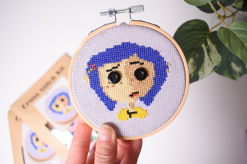 May include: A cross-stitch embroidery hoop with a purple and blue character with a yellow shirt and black button eyes. The character is on a light purple background.