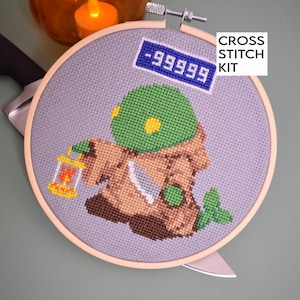 Puede incluir: Un kit de punto de cruz con un personaje verde y marrón que sostiene una linterna. El personaje lleva una túnica marrón y un sombrero verde. El kit incluye un cartel azul con el texto " -99999" y el texto "CROSS STITCH KIT".