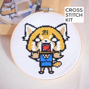 May include: A cross stitch kit featuring a pixelated image of a red panda with a blue shirt and the text "!!" on its head. The kit is in a round wooden embroidery hoop.