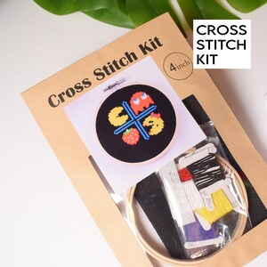 May include: A brown cardboard box with a clear plastic window showing a 4-inch embroidery hoop with a black fabric and a cross stitch design of Pac-Man, a ghost, a strawberry, and a yellow Pac-Man. The box also includes a variety of colored embroidery floss and a needle. The box has the text "Cross Stitch Kit" and "4 inch" printed on it.
