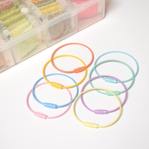 May include: A collection of colorful, circular keychains in shades of orange, yellow, blue, pink, green, and purple. Each keychain has a matching clasp. A clear plastic organizer filled with spools of thread is in the background.