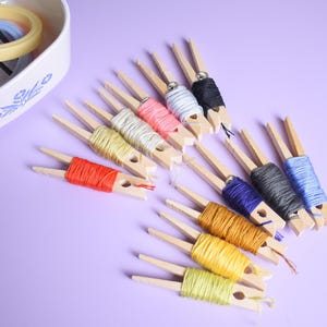 May include: A collection of wooden clothespins wrapped with various colors of embroidery floss, including red, yellow, gold, orange, blue, grey, black, pink, and white. A white bowl containing small embroidery hoops is visible in the upper left corner.