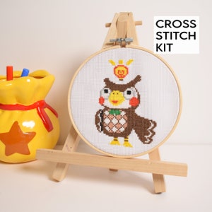 May include: A cross stitch kit featuring a brown owl with a yellow beak and large eyes. The owl is wearing a crown with a red star and is holding a green and white pineapple. The kit is displayed on a wooden easel with the text "CROSS STITCH KIT" above it.