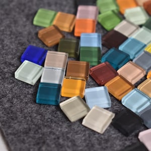 May include: A collection of small, square glass tiles in various colors, including blue, green, orange, and brown. The tiles are scattered on a gray felt surface, creating a mosaic effect. These tiles are ideal for craft projects.