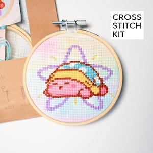 May include: A cross stitch kit featuring a cartoon character sleeping on a star. The design is stitched on a light blue, pink, and yellow background. The kit includes a wooden embroidery hoop and a cardboard backing. The text "CROSS STITCH KIT" is visible.