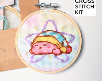 May include: A cross stitch kit featuring a cartoon character sleeping on a star. The design is stitched on a light blue, pink, and yellow background. The kit includes a wooden embroidery hoop and a cardboard backing. The text "CROSS STITCH KIT" is visible.