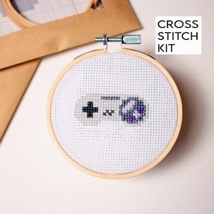 Puede incluir: Un kit de punto de cruz con un mando de videojuego blanco y gris con un botón morado. El kit incluye un aro de bordado de madera y tela Aida blanca.