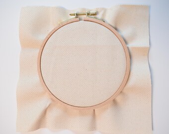 May include: A wooden embroidery hoop with a brass clasp holds a piece of beige cross-stitch fabric. The fabric is stretched taut within the hoop, ready for needlework. The background is a plain white surface.