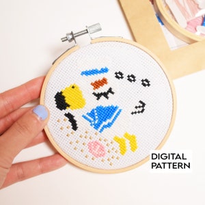 May include: A white embroidery hoop with a colorful cross-stitch pattern of a bird, a bow, and other geometric shapes. The text "DIGITAL PATTERN" is printed below the hoop.