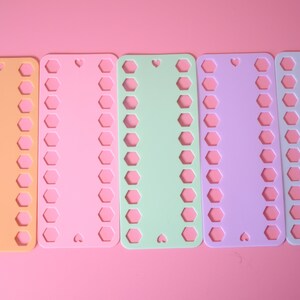 K&ouml;nnte beinhalten: F&uuml;nf farbige Kunststoff-Fadenorganisatoren in Orange, Rosa, Gr&uuml;n, Lila und Wei&szlig;. Jeder Organizer hat eine Reihe sechseckiger L&ouml;cher und einen herzf&ouml;rmigen Ausschnitt. Der Hintergrund ist einfarbig rosa.