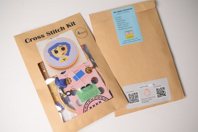 May include: A brown paper envelope with a clear plastic window showing a cross stitch kit. The kit includes a 4-inch embroidery hoop, Aida fabric, DMC threads, a needle, and a design chart. The design is a cartoon character with blue hair and a yellow shirt. The envelope also has a label with the text "DIY Cross Stitch Kit" and a QR code.